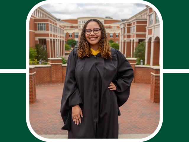 Carmen Rudd poses with her left hand on her hip and the right hand to her side, she wears a black grad gown. Her hair is curly and light brown. She smiles in front of the student union at unc charlotte 