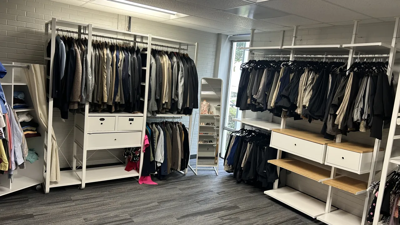 A neatly organized professional clothing closet with racks of business attire, including blazers, dress shirts, slacks, and skirts arranged by color and style. The space features white shelving units with drawers, a full-length mirror, and a display of dress shoes in the background. The room has modern carpet and natural light coming through a large window.
