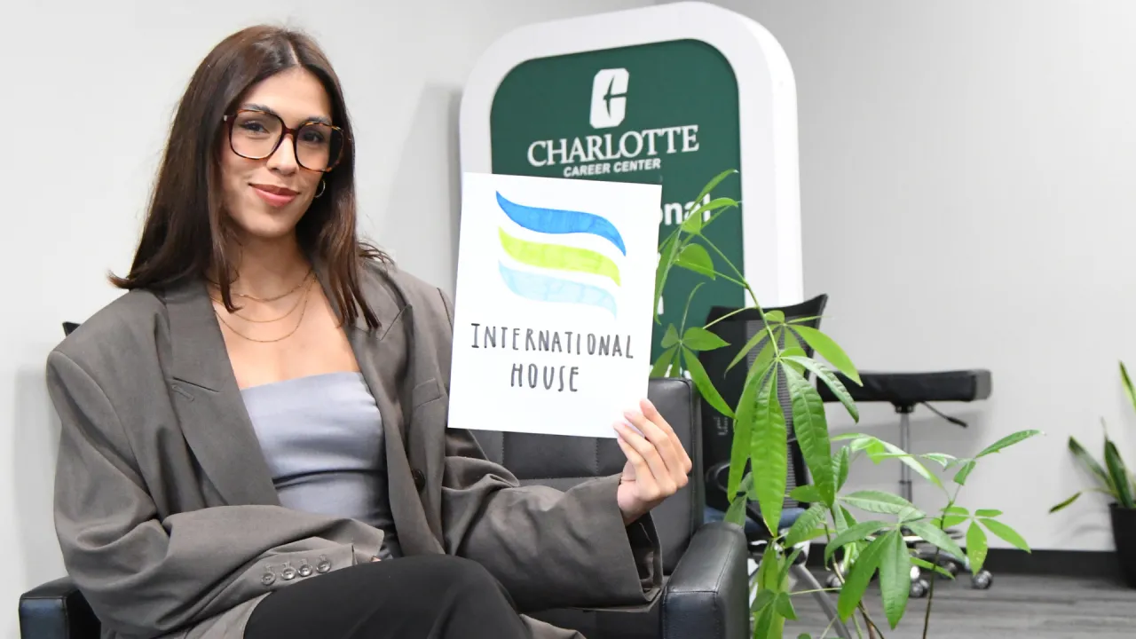 Sara Guerra Burgos sits in the UNC Charlotte Career Center in professional clothing while holding up a hand-made sign that has the International House logo on it. She smiles studiously.