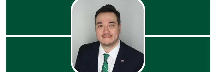 Chris Brasel is in focus wearing a black suit and green tie, he looks poised with a small, faint smile, looking at camera. Short brown hair and brown eyes. He is before a neutral, gray background.