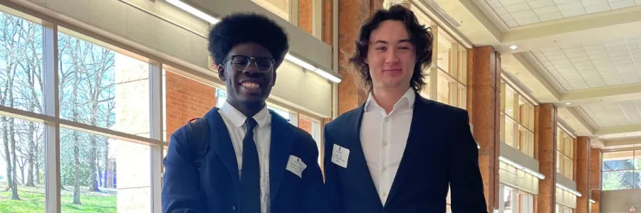 Two people stand next to each other wearing suits and one is wearing a tie