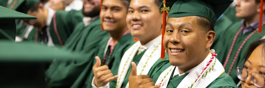 A group of niners pose in the audience at graduation, two of them are holding up the pick axe hand symbol but the first person is in full focus