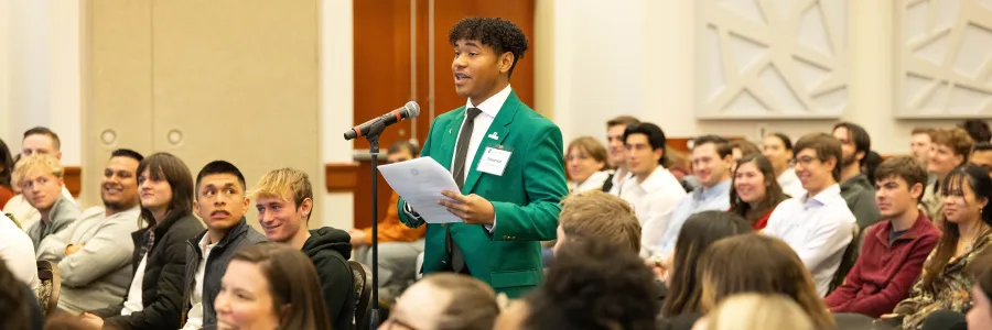 A UNC Charlotte student stands in the middle of a full audience, behind a microphone, speaking from a piece of paper while the audience appears to be reacting to something with humor