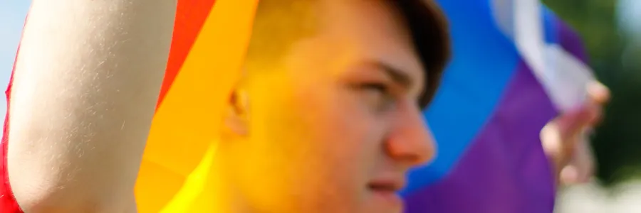 Profile angle of white person holding LGBTQ flag, with the colors reflecting off their face through it