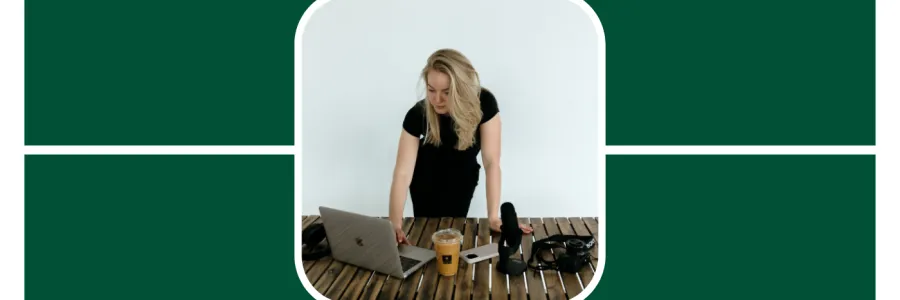 Madison Sims stands over a wooden table looking down on her laptop which is next to a coffee cup, camera, and podcast equipment. 