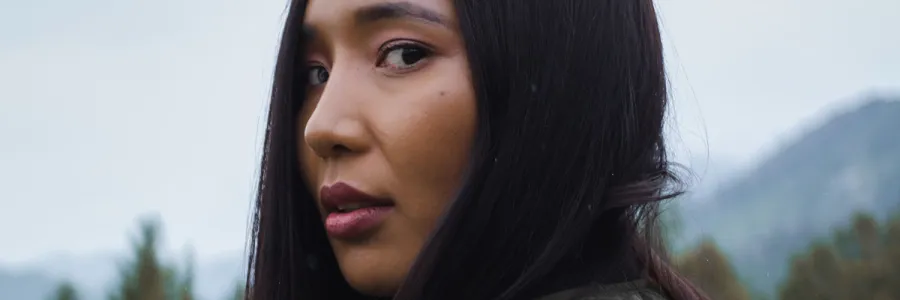 Native American woman with straight black hair with head turned so she's looking slightly towards the camera with mountains in distance