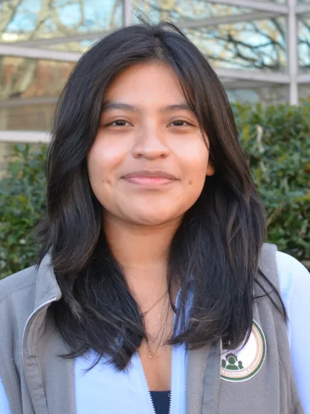 Ali smiles, wearing a vest, with a blurred green background.
