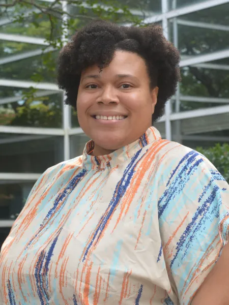 Alisha stands with a turned shoulder, wears a brightly colored shirt, smiles, and the background is blurred