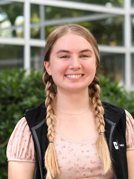 Baleigh smiles at the camera wearing a black vest and pink top, the background is greenery that is blurred