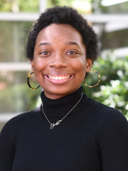 Cydnei wears a black turtleneck and brown hoop earrings. She has short, dark hair and smiles with her teeth. She poses in front of greenery