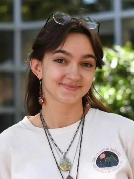 Lily is wearing a white tshirt and multiple layered necklaces, her hair is down and brown, she smiles slightly and her eye glasses are on top of her head.