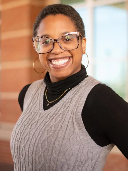 Rianna smiles brightly, wearing a black turtle neck, gold necklace, and a grey sweater vest.