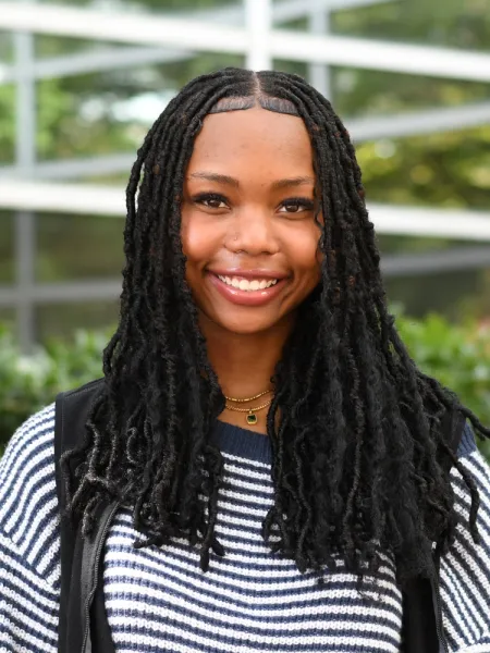 Theresa smiles at the camera, wearing a blue and white thin striped shirt, the background is blurred greenery