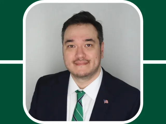 Chris Brasel is in focus wearing a black suit and green tie, he looks poised with a small, faint smile, looking at camera. Short brown hair and brown eyes. He is before a neutral, gray background.