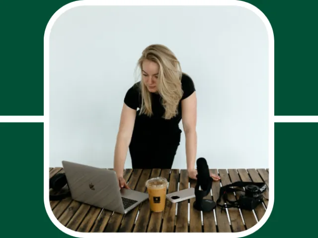 Madison Sims stands over a wooden table looking down on her laptop which is next to a coffee cup, camera, and podcast equipment. 