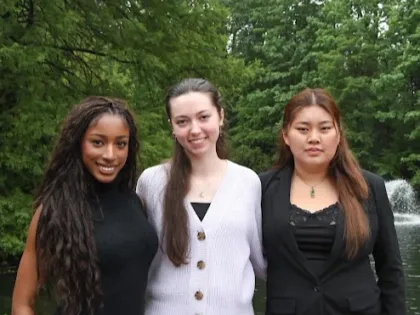 Jiya, Ryleigh, and Amelia pose together wearing professional level clothing. They smile. Background is a lake and filled with trees and greenery.