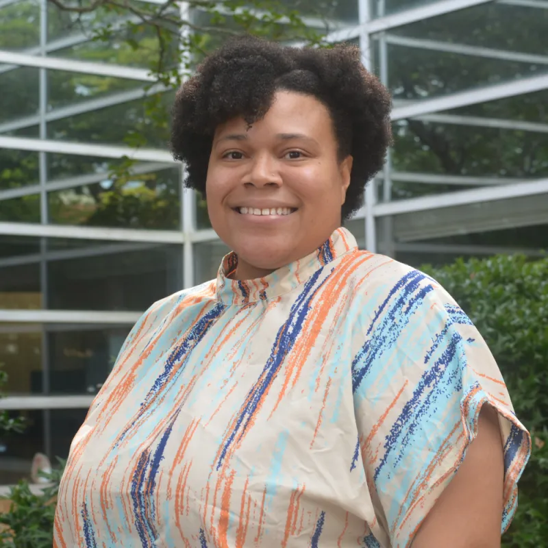 Alisha stands with a turned shoulder, wears a brightly colored shirt, smiles, and the background is blurred