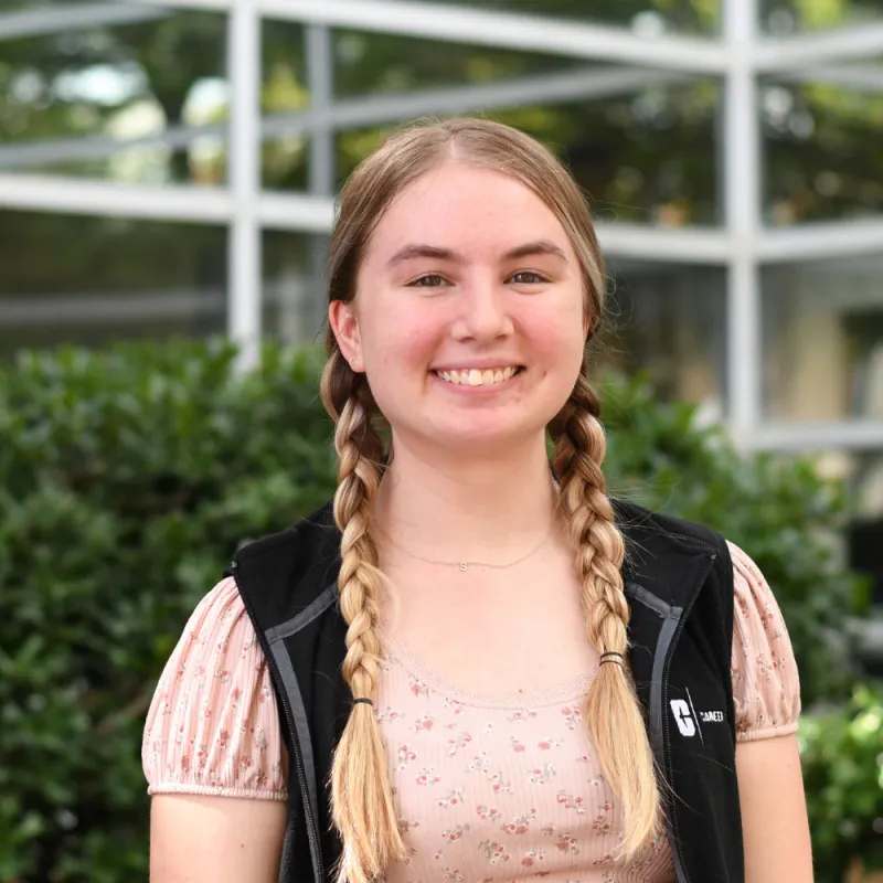 Baleigh smiles at the camera wearing a black vest and pink top, the background is greenery that is blurred