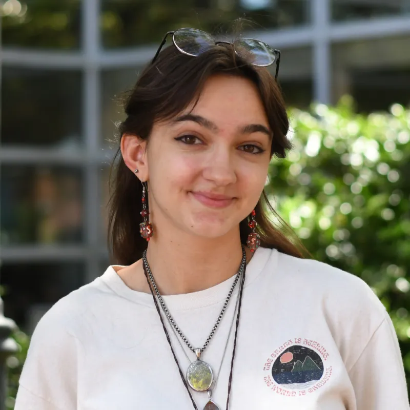 Lily is wearing a white tshirt and multiple layered necklaces, her hair is down and brown, she smiles slightly and her eye glasses are on top of her head.