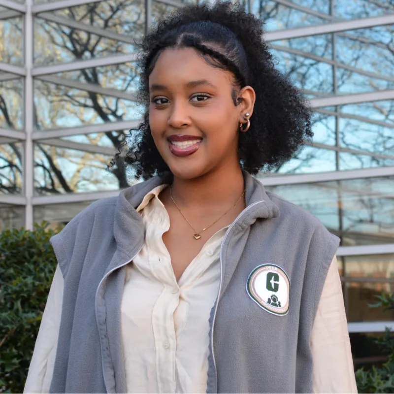 Mary smiles brightly, wears a grey vest, standing in front of blurred modern windows.